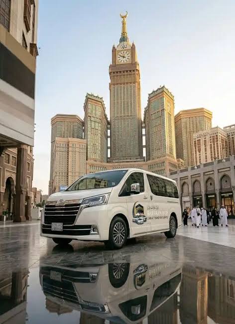 Van blanc devant la tour de l’horloge de Makkah, sur une chaussée mouillée qui reflète la lumière.