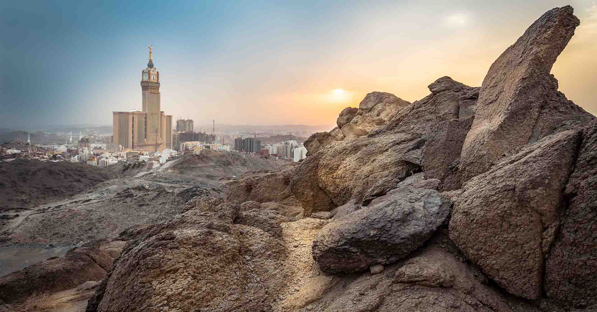Rochers au premier plan et skyline de Makkah au coucher du soleil, la tour de l’horloge visible au loin.